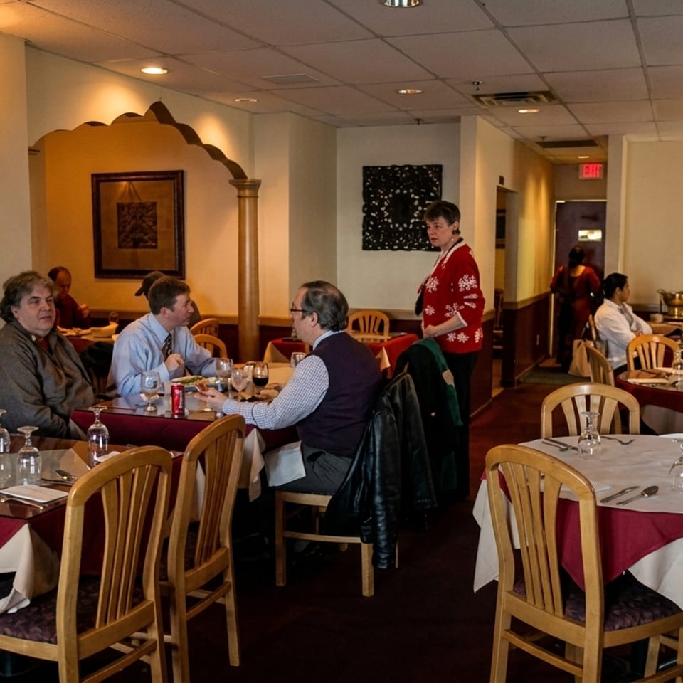 Warmly lit restaurant dining room with white and burgundy tablecloths, seated diners, and a server in a red sweater.