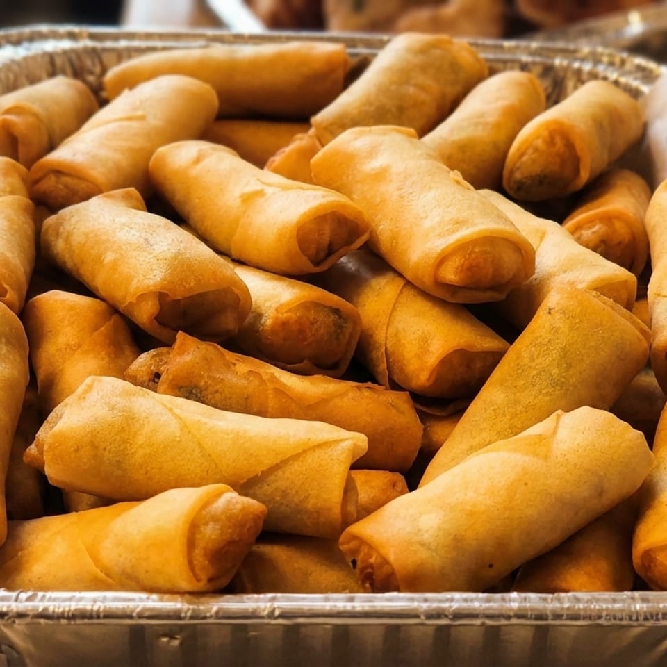 Aluminum tray filled with golden fried spring rolls and crispy triangular pastries on a restaurant counter.