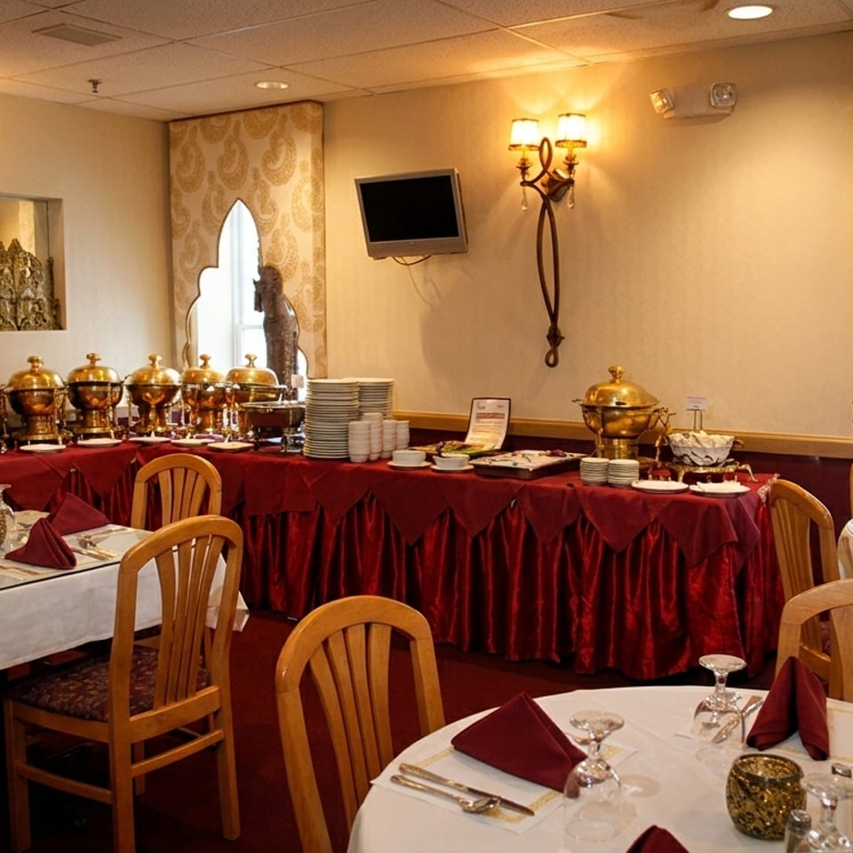Elegant restaurant dining room with white-clothed tables, red napkins, buffet chafing dishes, ornate décor, and statues.